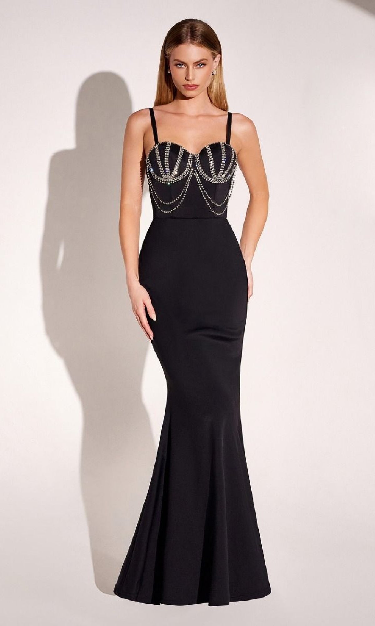 A woman wears the Amoris T9003 Beaded-Bustier Long Prom Gown—a fitted black mermaid dress with thin straps and decorative silver chains—standing against a plain light background.